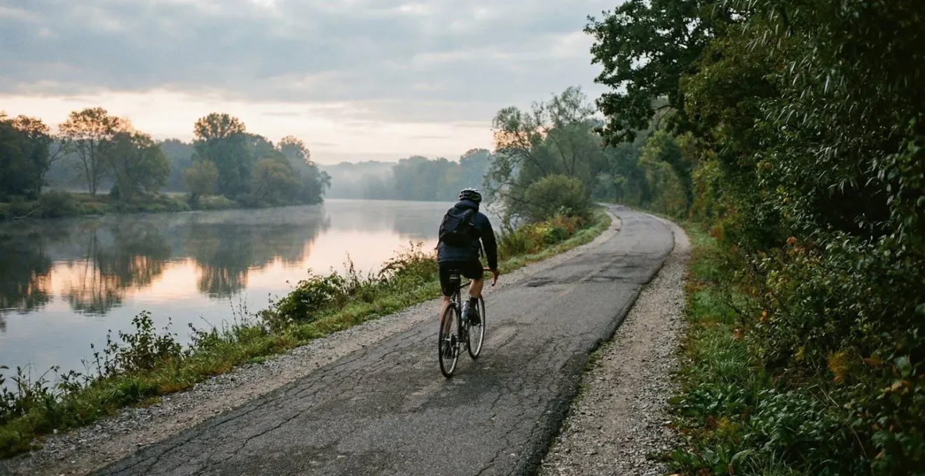 Un cycliste roule de dos sur une piste cyclable longeant la Marne, lumière douce du matin filtrant à travers les arbres